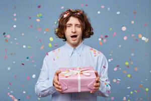 Man holding a decorated gift in a big box, presents it, winkles, in a playful mood, confetti falling down on a blue background. photo