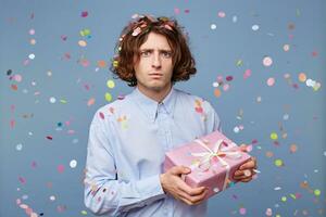 Puzzled, frightened guy posing with a decorative pink gift box looks as if he is afraid that someone will take it away, holds present tightly in his hands, against the background of falling confetti. photo