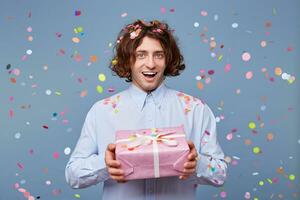 Blithesome long hair guy enjoying birthday with presents. Indoor shot of carefree european man in red shirt having fun at party photo