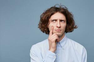 Portrait of focused smart and creative male with long messy hair, holding fingers on cheek and chin, gazing left with thoughtful expression, thinking over blue background photo