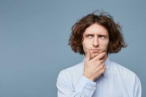 Confident pensive handsome young man doubts, touching face while thinking is looking away wearing white shirt, standing on the blue background. Empty copy space for promotion photo