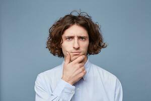 Portrait of a pensive young man dressed in shirt holding hand on his chin isolated over blue background. Creative and artistic male gazing thoughtfully at camera, thinking or making up ideas photo