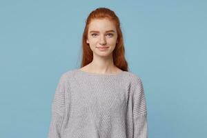 Young attractive beautiful pretty red-haired girl with long hair looks into the camera, smiles sweetly, is dressed in an oversized simple warm sweater, isolated on a blue background photo