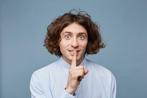 Cute young man having secret, holding finger on lips and showing silence sign, standing on blue background.Emotional male with long messy hair, ask to keep confidential information in secret photo