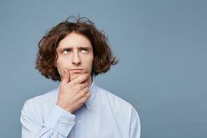 Portrait of suspicious pensive young male in casual shirt touching face while thinking over something, trying to come up with solution, having perplexed and puzzled expression over blue background photo