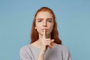 Portrait of red-haired concerned girl demonstrating a gesture of silence,keeps finger at her mouth, looks at the camera, asks to keep a secret, secrecy, information confidential, over blue background. photo