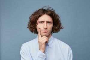 Thoughts and doubts concept. Man with long messy hair thoughtful face, holding fingers on cheek and chin, gazing camera. Guy with untidy hairstyle has doubts, copy space, blue background. photo