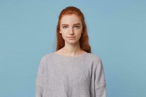 Young beautiful girl with long red hair and blue eyes with freckles looks straight into the camera feels calm and confidently isolated on a blue background photo