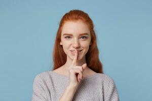 Headshot of attractive cheerful red-haired young female depicts a gesture of silence,keeps finger at her mouth,playfully looks at the camera, smiles,asks to keep a secret,privacy,over blue background. photo