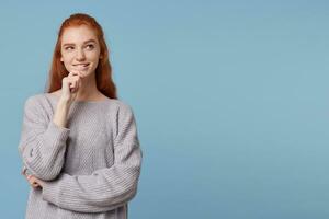 Red-haired beautiful girl stands holding her hand near the chin slightly bit her lip looking aside at the copy space, dreams, plots, plans, ponders ideas, cheerful playful mysterious look photo