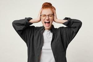 A young red-haired woman stands with eyes closed, her hands on her head depicts a loud strong cry isolated on white background. photo