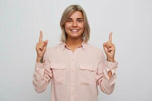 Closeup of cheerful attractive blonde young woman with braces on teeth wears pink shirt looks happy and points up by fingers of both hands isolated over white background photo