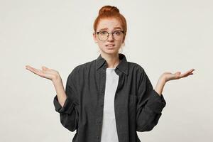 Studio photo of a girl with red hair wearing glasses dressed in a man's black shirt depicts helplessness hopelessness holding hands to the sides with her palms upwards. Confused scared apologizes.