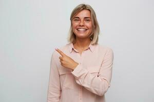 Closeup of cheerful beautiful young woman with blonde hair and braces on teeth wears pink shirt looks happy and points to the side by finger isolated over white background photo