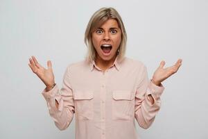 Closeup of mad shocked young woman with blonde hair, opened mouth and braces on teeth wears pink shirt feels angry, shouting and holding copy space on both palms isolated over white background photo