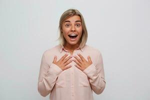 Portrait of surprised excited blonde young woman with opened mouth and braces on teeth wears pink shirt looks amazed and feels happy isolated over white background photo