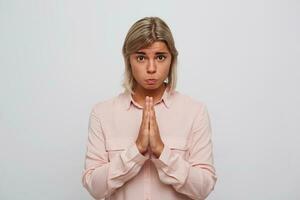 Portrait of sad cute woman with blonde hair wears pink shirt feels upset and keeps hands in praying position isolated over white background Looks directly in camera photo