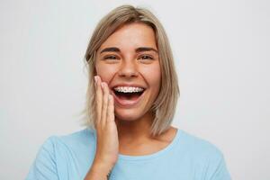 Cheerful beautiful young woman with blonde hair and braces on teeth laughing and touching her face by hand isolated over white background photo