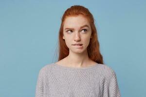 Portrait of a young red-haired teen girl looks away bit her lower lip, doubts, thinks over options, is unsure, plagued by doubts, isolated on blue background photo