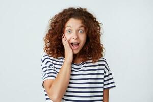 Portrait of surprised excited cute young woman with curly hair and opened mouth wears striped t shirt looks amazed and shouting isolated over white background photo