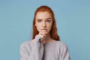 Portrait of a girl with red hair, looks camera holds her hand near the chin bit her lip, reflects on something, feels a little hesitant wants to make some decision, doubts, isolated on blue background photo