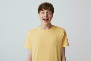 Portrait of surprised attractive young man with short haircut and braces on teeth wears yellow t shirt standing with opened mouth and feels excited isolated over white background photo