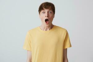 Closeup of astonished handsome young man with short haircut and opened mouth wears yellow t shirt shouting and looks shocked isolated over white background photo