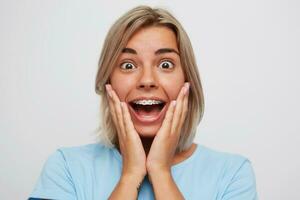 Portrait of happy surprised young woman with blonde hair and braces on teeth looks excited and touching her cheeks by hands isolated over white background photo