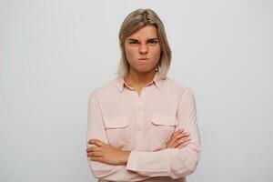 retrato de irritado ceñudo rubia joven mujer usa rosado camisa mantiene brazos cruzado y haciendo enojado cara aislado terminado blanco antecedentes foto