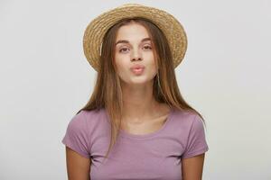 Close up of cute lovely girl in casual outfit in a straw hat with a pink ribbon sending blowing kiss with pout lips looking at camera isolated on white background. Affection feelings concept photo