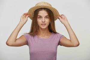 Charming young female keeps in hands straw hat, tries it on or corrects, imagines a vacation by the sea, hot summer, dreams of summer trip, against white studio background photo