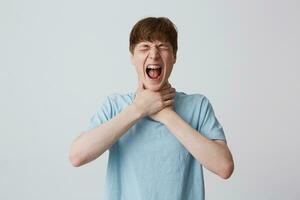 Portrait of crazy desperate young man student with closed eyes and braces on teeth wears blue t shirt strangles himself with both hands and screaming isolated over white background photo