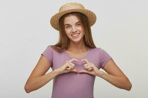 Romantic portrait of tender charming woman on a white background. Girl shows heart shape gesture with hands. Female in a straw hat with a pink ribbon showing love symbol, looking at camera photo