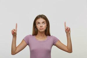 Impressed girl feels unhappy,shows with forefingers of both hands up to the copy space, corners of the lips down, upset, being displeased see some item, isolated over white background. photo