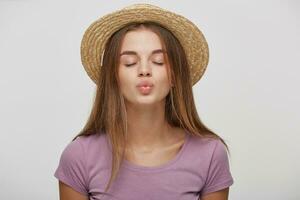 Portrait of cute lovely girl in casual outfit in a straw hat with a pink ribbon sending blowing kiss with pout lips with eyes closed, isolated on white background. Affection feelings concept photo