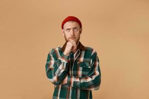 Forgetful confused curious bearded man in checkered shirt and hat, holding fist on chin and with thoughtful clueless expression, looking right upper corner, isolated over beige background. photo