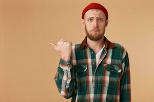 An upset guy with a sad look in his eyes with a displeased facial expression shows his thumb on his left hand on the empty space for promotion. photo