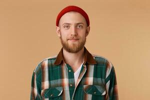 atractivo joven hombre en estudio mirando a cámara. retrato de un normal chico con ancho en rojo sombrero y a cuadros camisa en frente de beige antecedentes foto