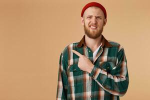 Unhappy bearded male in red hat without pleasure points with index finger at left corner, shows copy space for advertisement or promotional text isolated over beige background photo