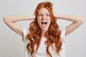 Closeup of crazy mad redhead young woman with long hair, freckles and opened mouth wears t shirt keeps hands on head and screaming over white background photo