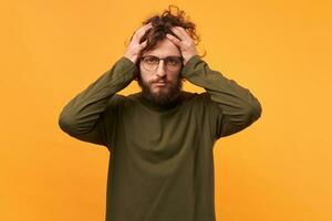 A man in glasses with a beard clutched at his head looks upset sad, got in desperate situations. Doomed exhausted dressed in ordinary clothes, isolated on a yellow background. photo