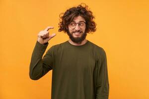 Handsome unshaven male with dark curly hair and thick bristle, shows something tiny with hands, dressed casually, isolated over yellow background. Young man demonstrates small thing indoor. photo