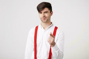 Attractive dark-haired unshaven guy wearing a white shirt and red suspenders shows his index finger at the camera as if he wants to say You are next, you get well done, you will succeed. photo