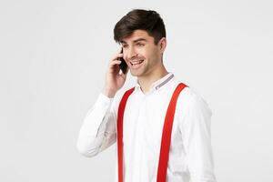 atractivo joven chico hablando en el teléfono mira alegre feliz, sonrisas siente felicidad abrumado con positivo emociones, genial noticias, victoria, usa camisa y rojo tirantes, en blanco antecedentes foto