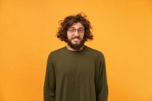 A man with a beard in glasses and curly dark hair looking at the camera, bite lip, confused bewildered as if made mistake, feel guilty isolated on a yellow background dressed in ordinary clothes. photo