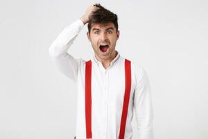 Indoor photo of stupefied astonished man wears shirt and red suspender, keeps hand on head, clutched head, says Omg, I am tired of that. Shocked male realizes his fault.