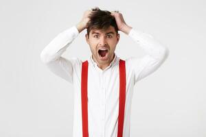 Man in panic clutched his hands in the hair on his head, wears shirt and red suspender, isolated over white background. Shocked male realizes his fault. photo