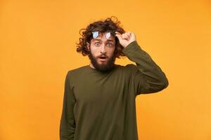 Student saw something unusual, curious, incredible, and even raised his glasses. Bearded curly-haired man raised his glasses looking at the camera his mouth opened in surprise photo