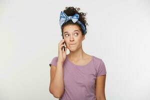 Girl is talking on the phone, looking to the side, bit her lower lip, pondering something, guilty and thinking how to explain it to her interlocutor, dreaming about something, over white background photo