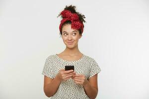 young girl with curly hair, looks happy like dreaming thinking on something pleasant and inspiring, eyes look aside, keeps smart phone in hands, dressed white t-shirt with dots, white background photo